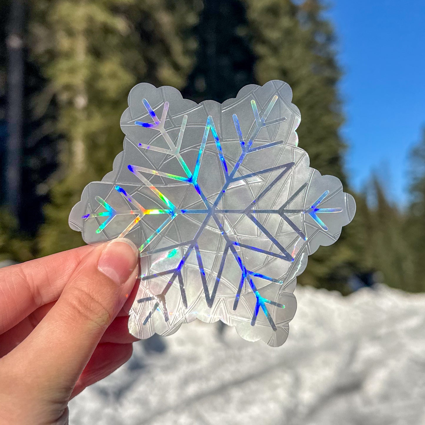 Holographic cozy winter snowflake suncatcher cling decal sticker. Held up in front of snow and trees.