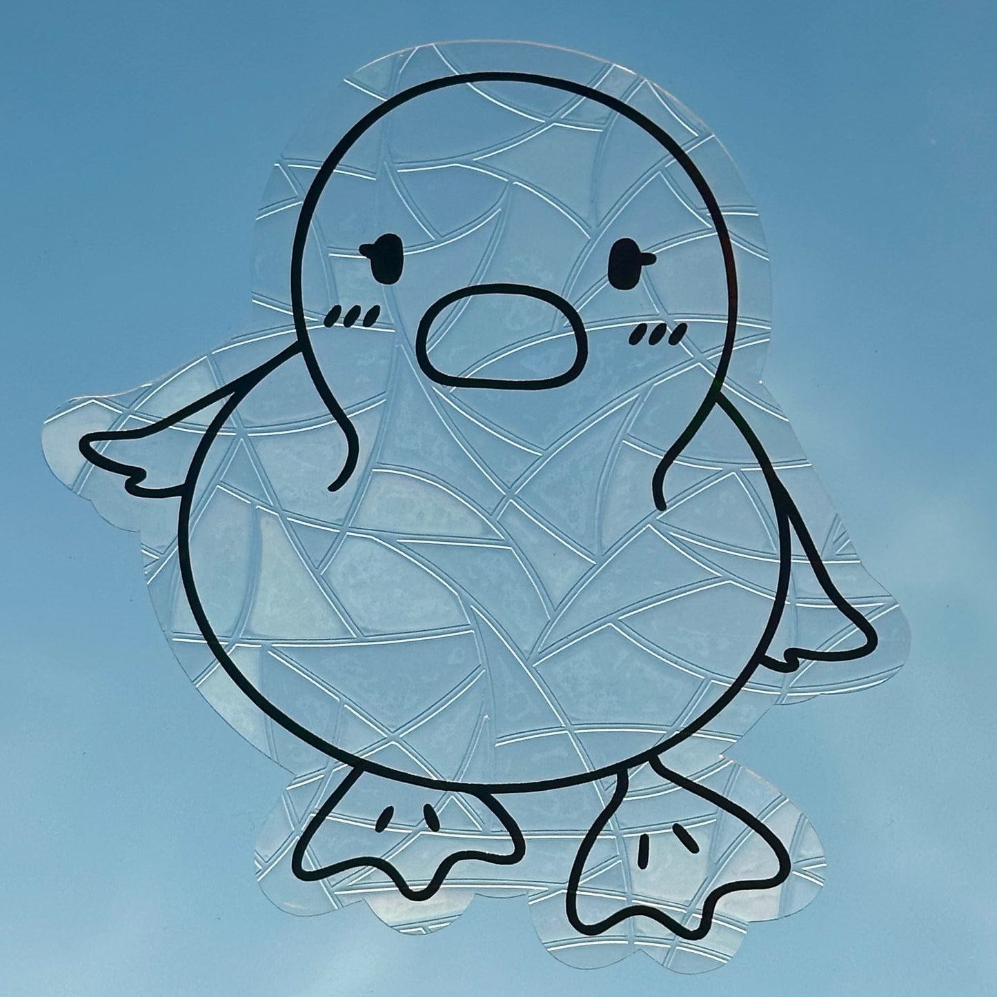 A Sun Catcher decal on glass in front of a blue sky.
The design features a baby duckling. The design is shiny and holographic, and sits on top of the clear rainbow-making film.