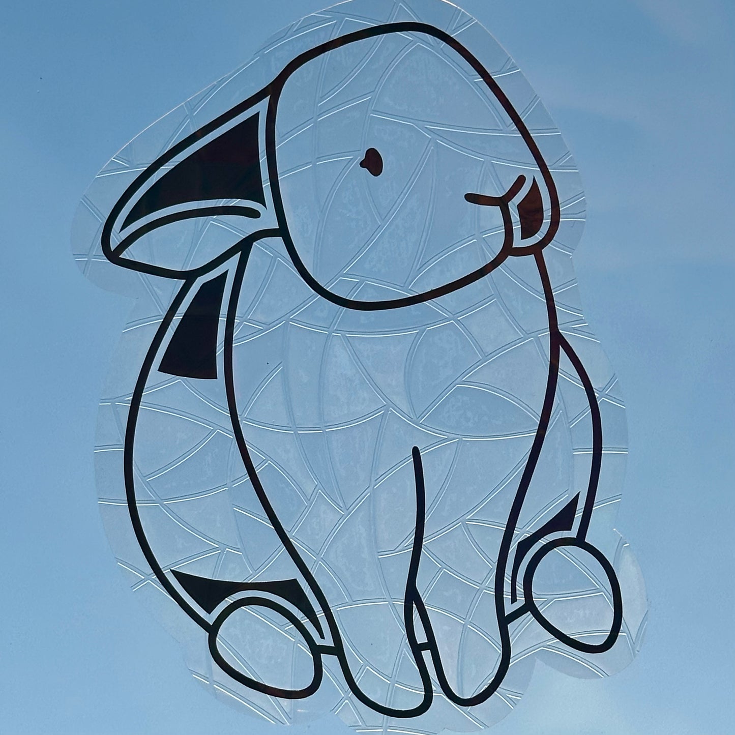 A Sun Catcher decal on glass in front of a blue sky.
The design features a sitting bunny rabbit. The design is shiny and holographic, and sits on top of the clear rainbow-making film.