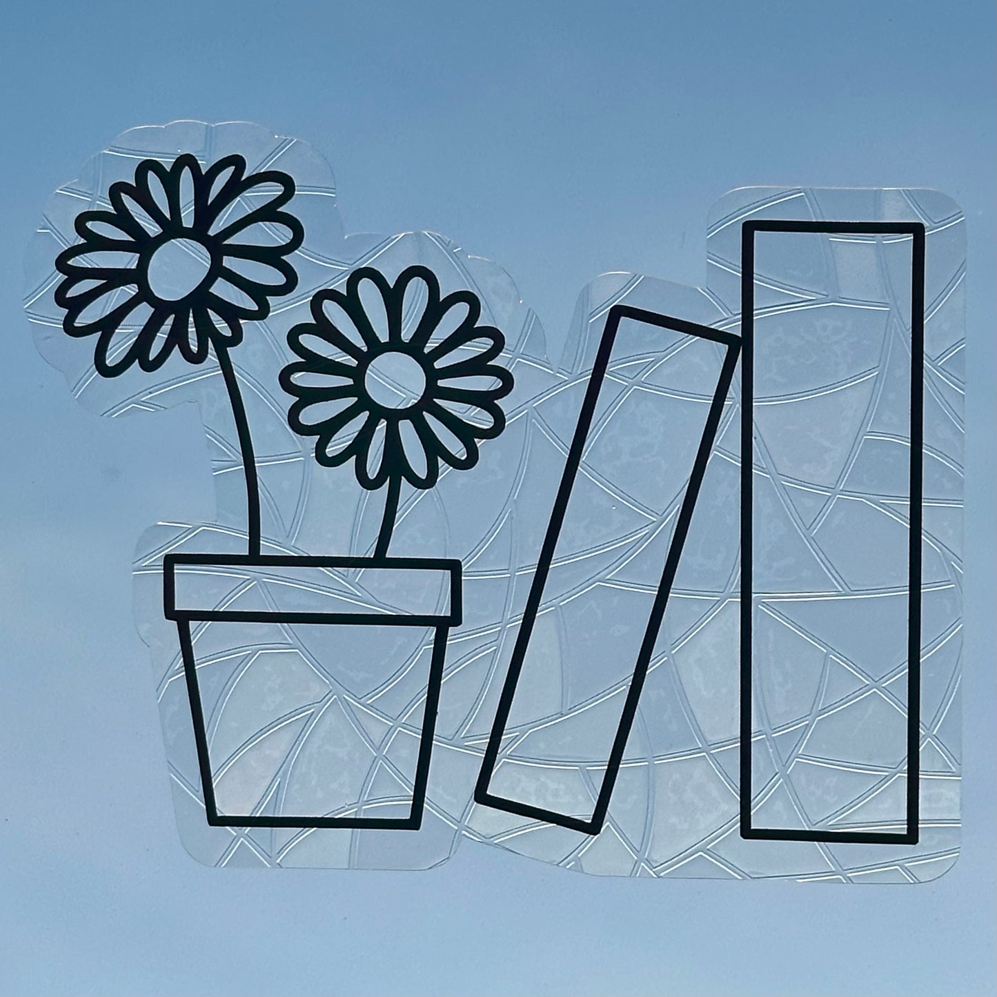A Sun Catcher decal on glass in front of a blue sky.
The design features a pot holding two daisies, and next to that, two books. The design is shiny and holographic, and sits on top of the clear rainbow-making film.