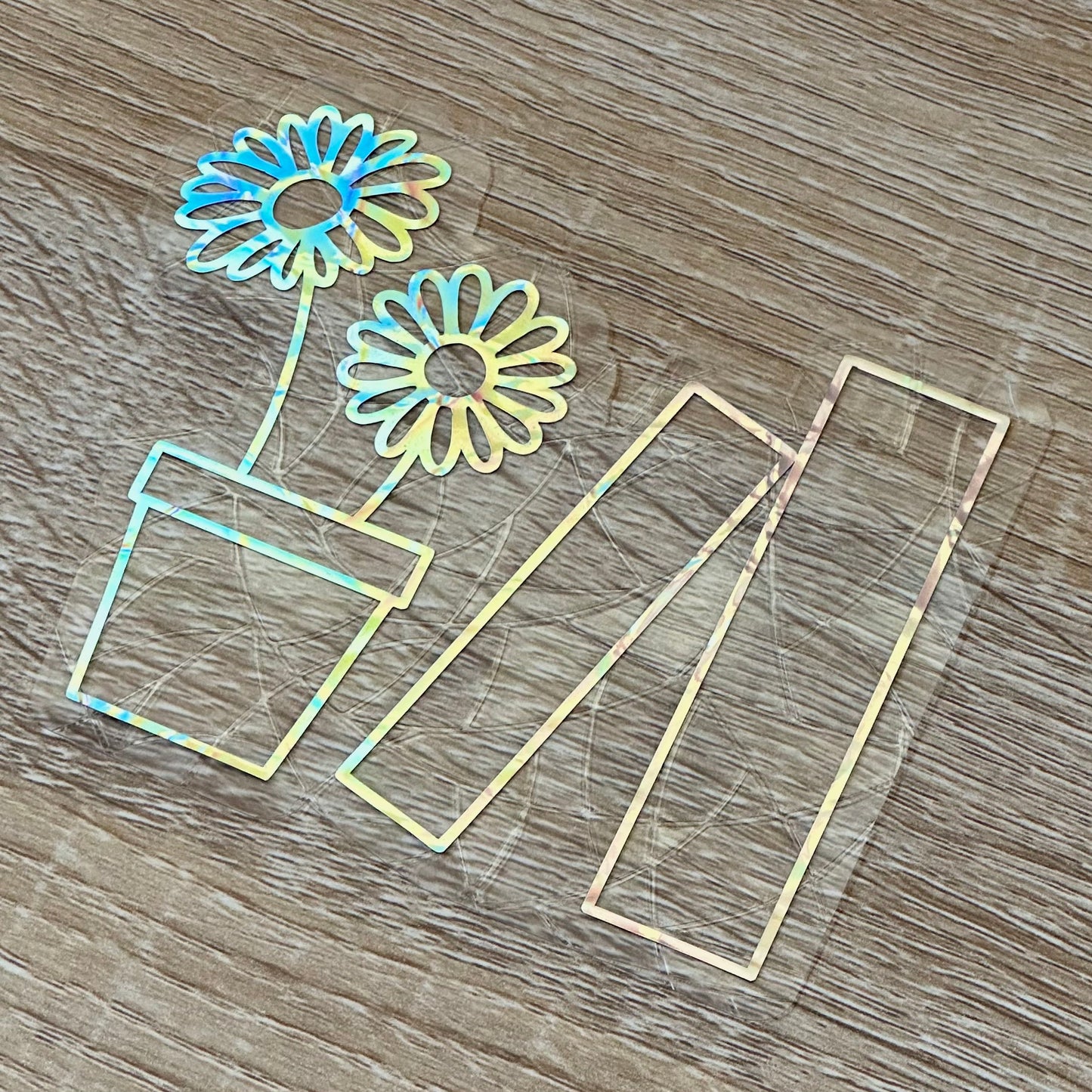 A Sun Catcher decal on a wooden desk top.
The design features a pot holding two daisies, and next to that, two books. The design is shiny and holographic, and sits on top of the clear rainbow-making film.
