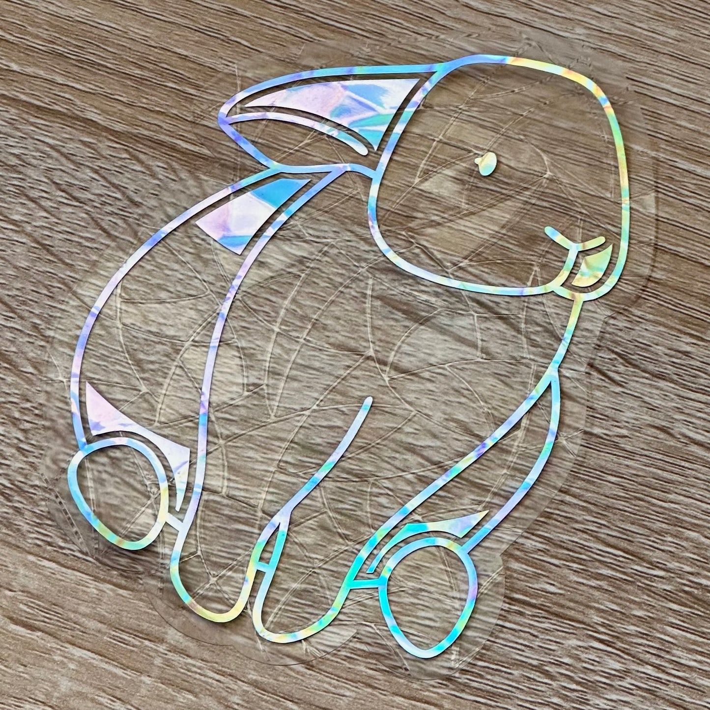 A Sun Catcher decal on a wooden desktop.
The design features a sitting bunny rabbit. The design is shiny and holographic, and sits on top of the clear rainbow-making film.