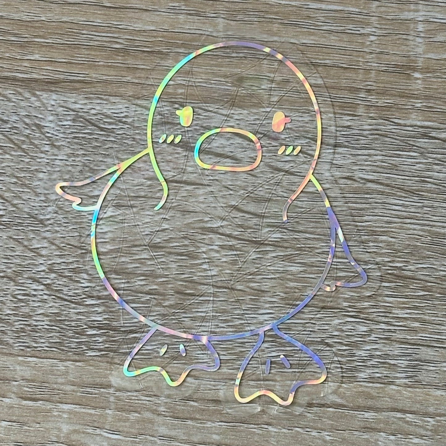 A Sun Catcher decal on a wooden desk top.
The design features a baby duckling. The design is shiny and holographic, and sits on top of the clear rainbow-making film.