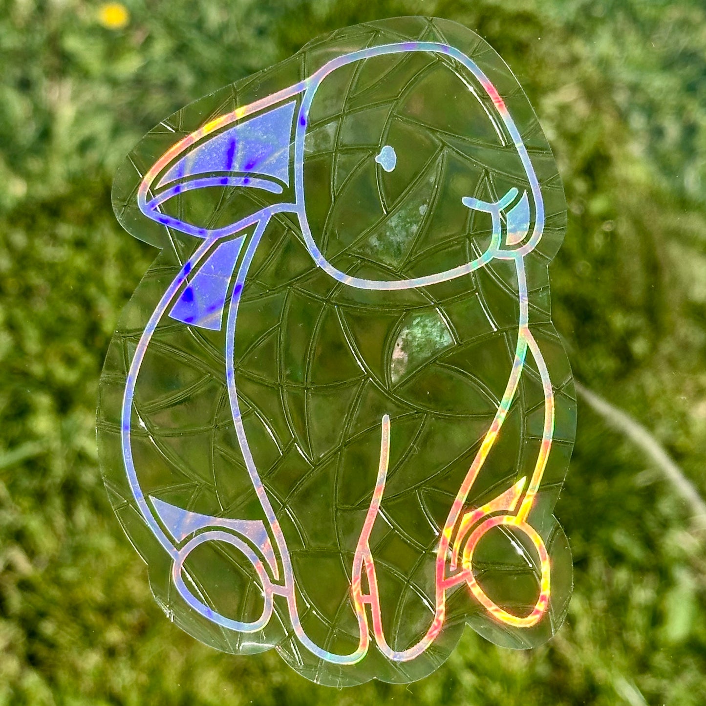 A Sun Catcher decal on glass in front of green grass.
The design features a sitting bunny rabbit. The design is shiny and holographic, and sits on top of the clear rainbow-making film.