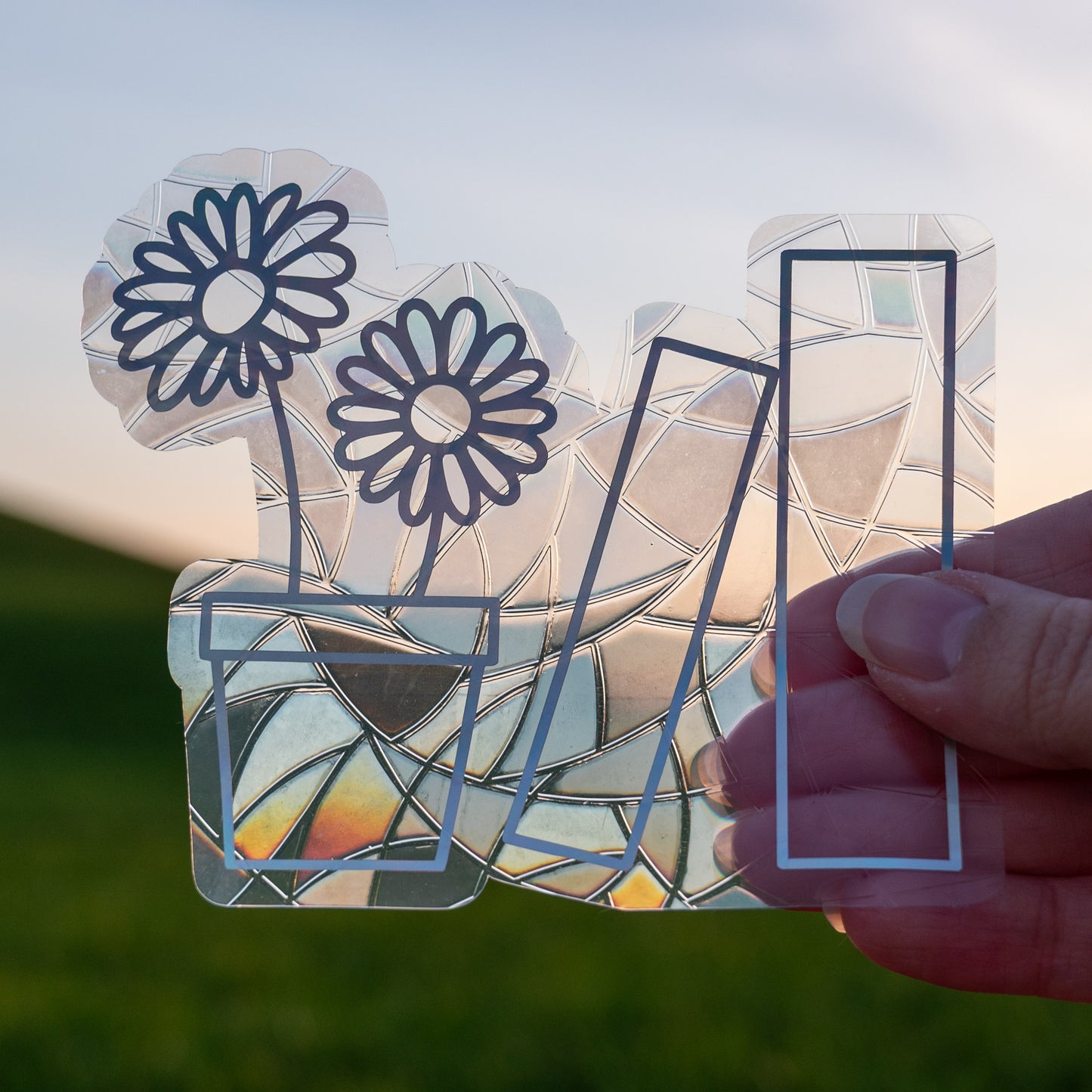A Sun Catcher decal held up in front of a blue sky and green hills.
The design features a pot holding two daisies, and next to that, two books. The design is shiny and holographic, and sits on top of the clear rainbow-making film.