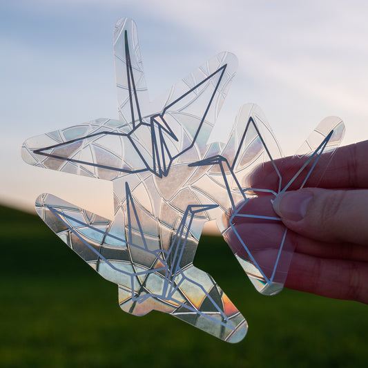 A Sun Catcher decal held up in front of a blue sky and green hills.
The design features three paper cranes. The design is shiny and holographic, and sits on top of the clear rainbow-making film.
