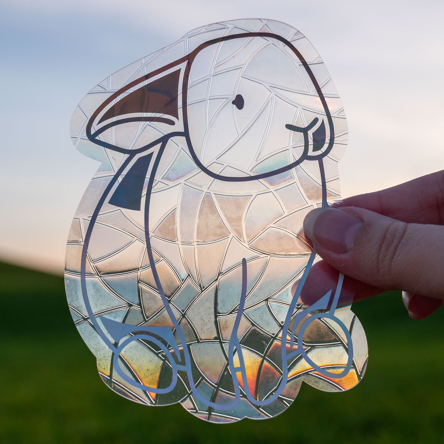 A Sun Catcher decal held up in front of a blue sky and green hills.
The design features a sitting bunny rabbit. The design is shiny and holographic, and sits on top of the clear rainbow-making film.