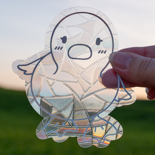 A Sun Catcher decal held up in front of a blue sky and green hills.
The design features a baby duckling. The design is shiny and holographic, and sits on top of the clear rainbow-making film.
