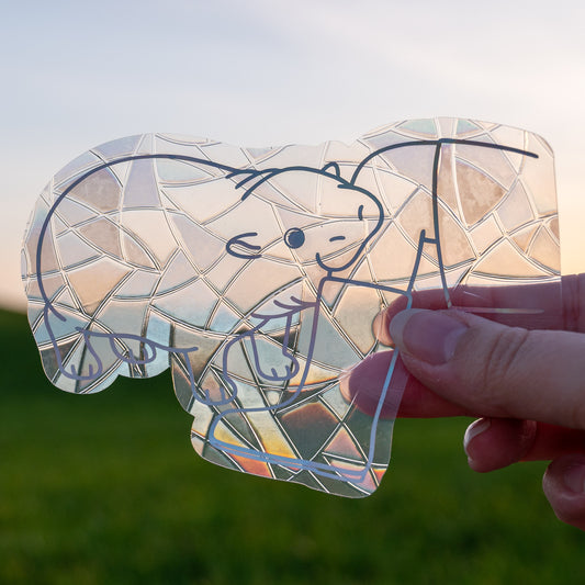 A Sun Catcher decal held up in front of a blue sky and green hills.
The design features a baby pygmy hippo biting a boot. The design is shiny and holographic, and sits on top of the clear rainbow-making film.