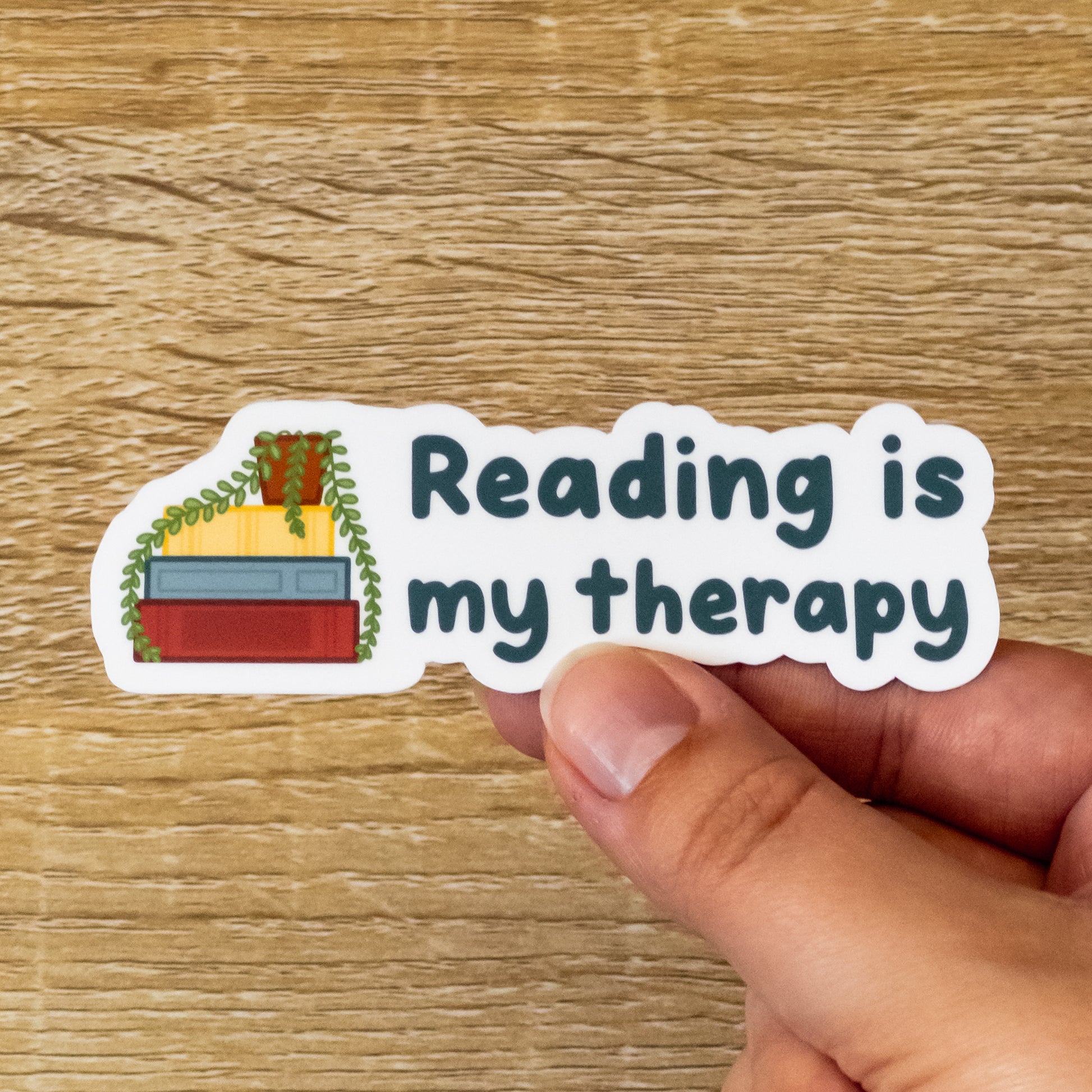 Glossy 3.6 inch wide sticker. Design is a stack of three books with a potted plant on top. The leaves drape over the red, yellow, and blue books. To the right, there is text that says, "Reading is My Therapy."