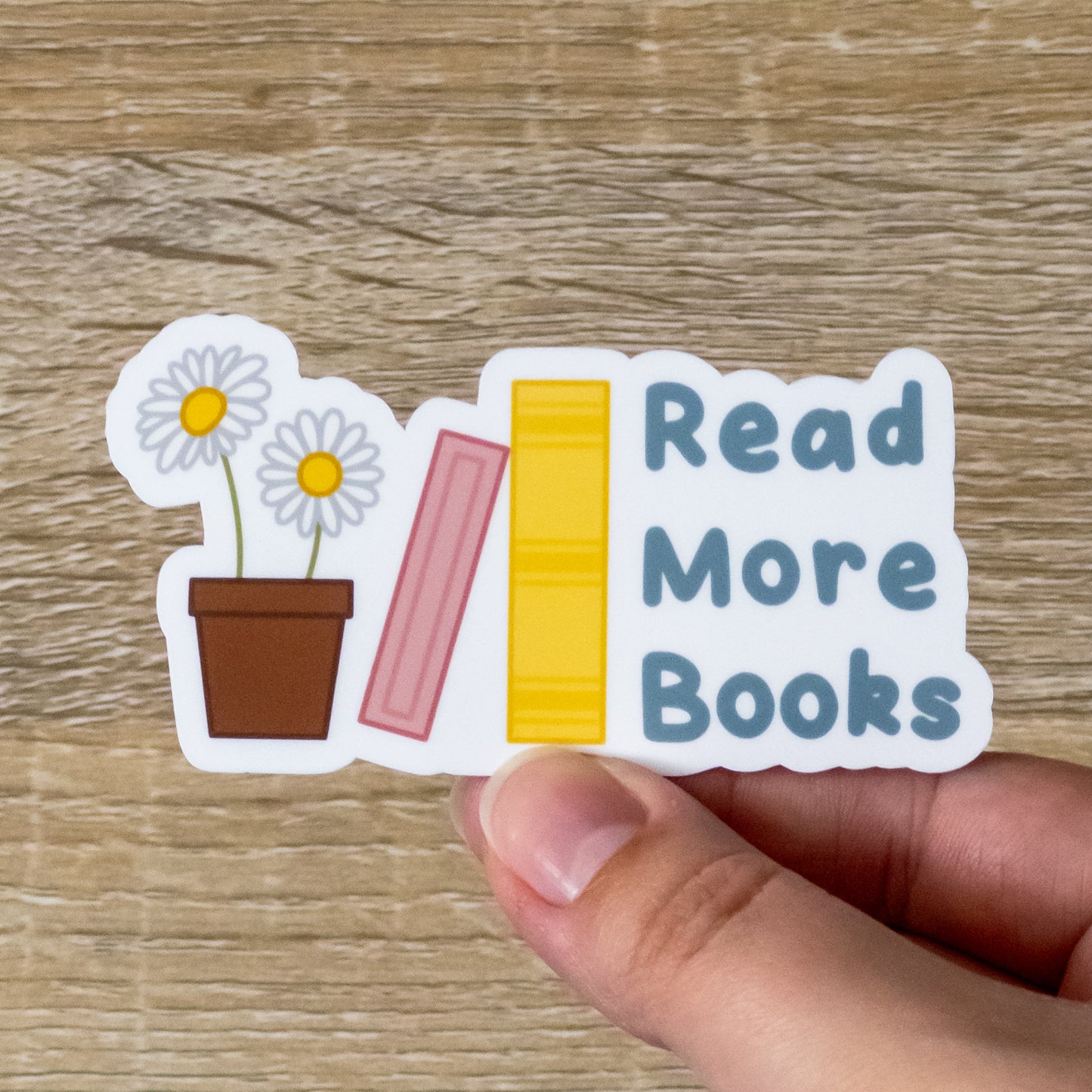 Glossy 3.4 inch tall sticker. Design is two daisies in a pot, a pink book leaning against a yellow book, and blue text that says, "Read More Books"