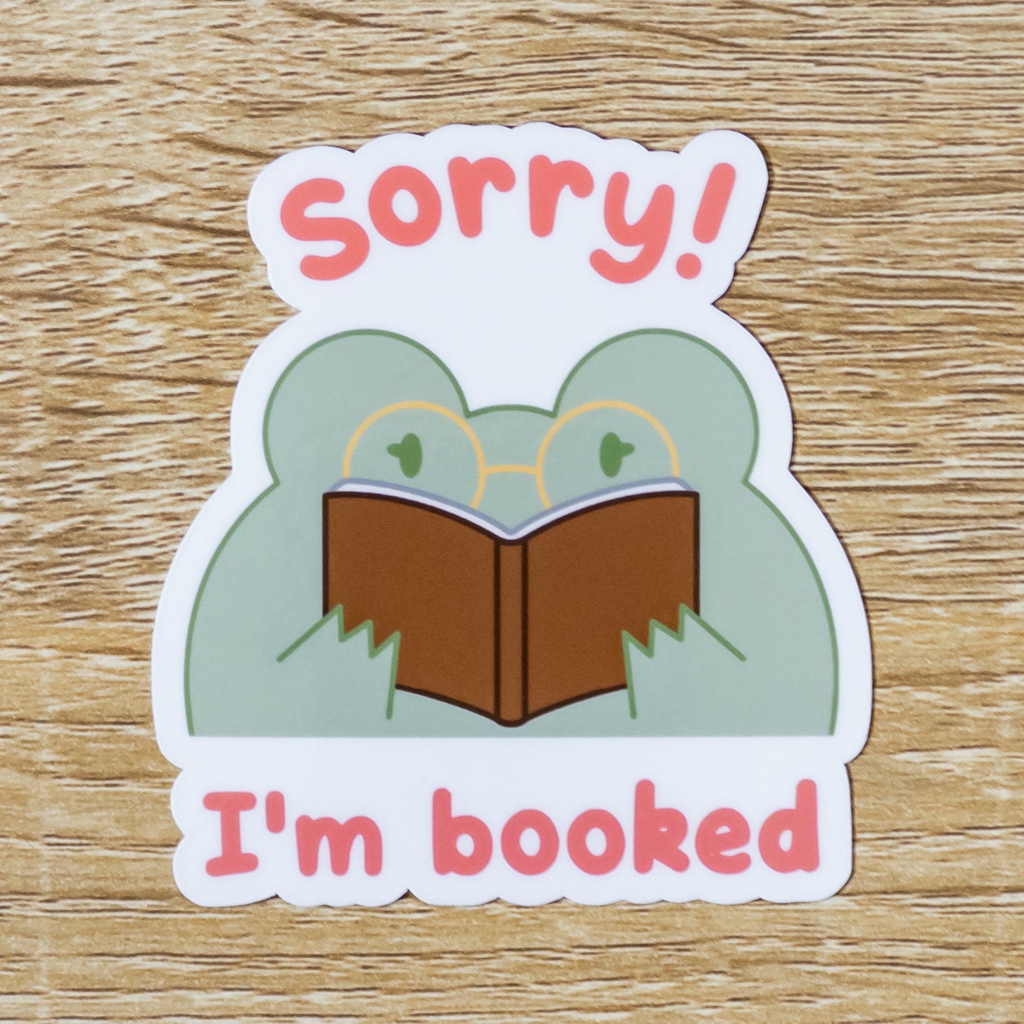 Glossy 3 inch tall sticker. Design is a green frog with gold glasses, reading a brown book, with the text, "Sorry! I'm Booked."