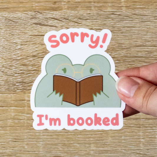 Glossy 3 inch tall sticker. Design is a green frog with gold glasses, reading a brown book, with the text, "Sorry! I'm Booked."