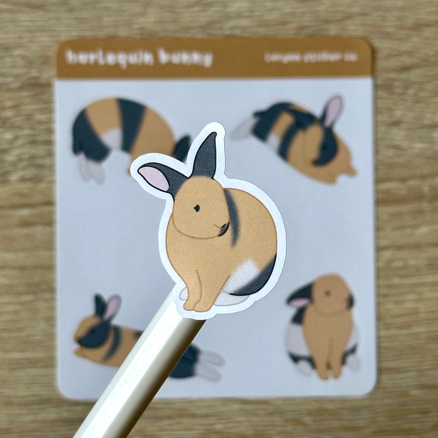 Holding up one sticker from the sheet. Sticker is an illustrated harlequin rabbit sitting and looking to the right. Rabbit is tan, black, and white. Sticker measures 1.2 x 1.5 inches. The four other stickers on the sheet are around that same size.