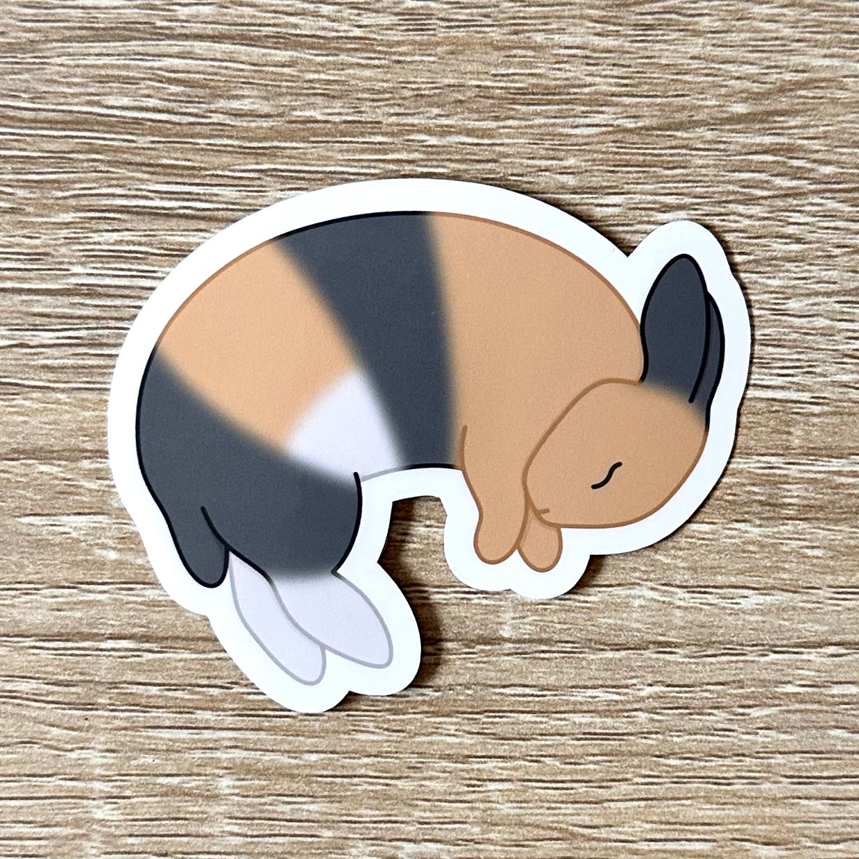 A sticker featuring an illustration of a harlequin bunny laying on his side, flopped and sleeping. The bunny is tan, white, and grey. Sticker is glossy and 2.7 x 2.1 inches.