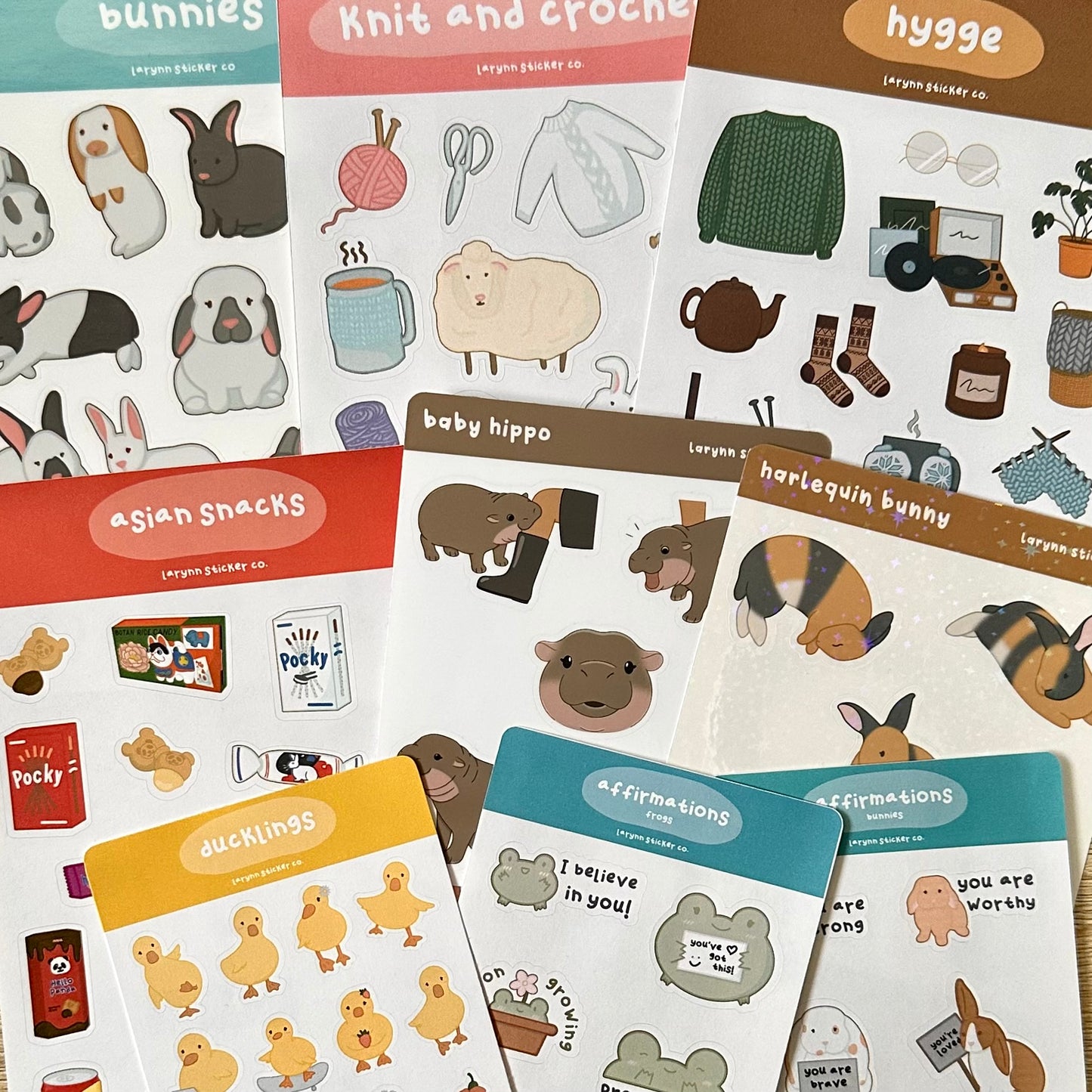 Various sticker sheet designs laying on a table. Includes matte, glossy, and holographic finishes. Designs include illustrated animals, foods, and other themes. Some sticker sheets are full sized, while others are mini.