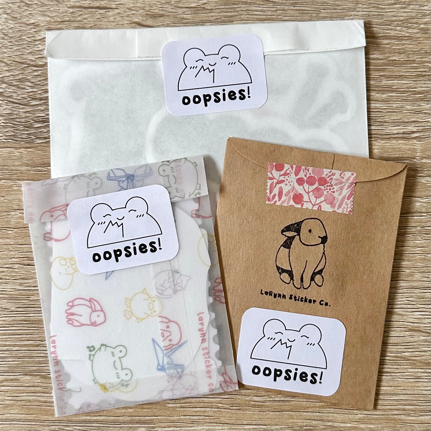 Three mystery packs of imperfect stickers, each one is in different packaging. The top is in a white glassine bag, the bottom left in themed vellum, and the bottom right a kraft paper envelope. Each one is labeled with an "oopsies" sticker