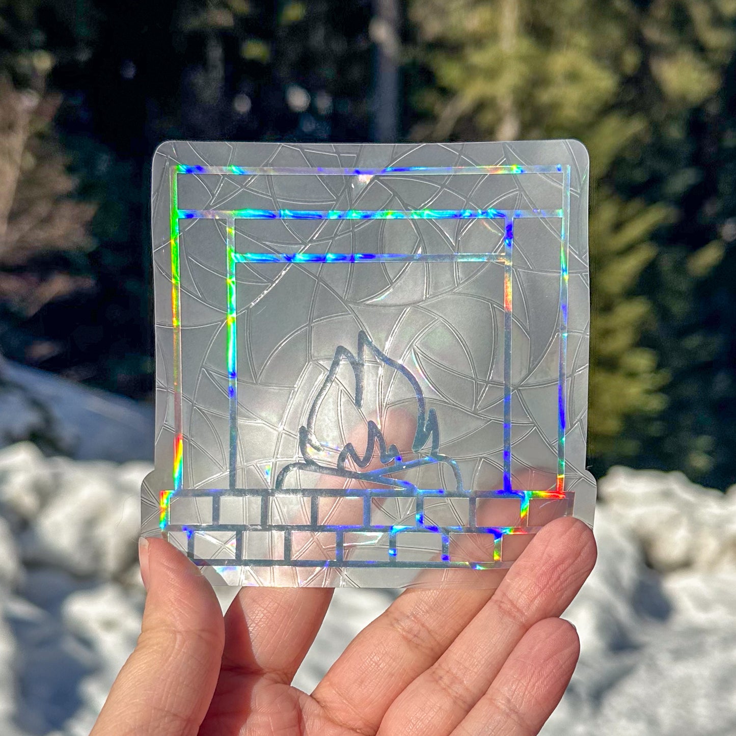 Holographic cozy winter fireplace suncatcher cling decal sticker. Holographic brick fireplace with two logs and a fire in the center. Held up in front of snow and trees.