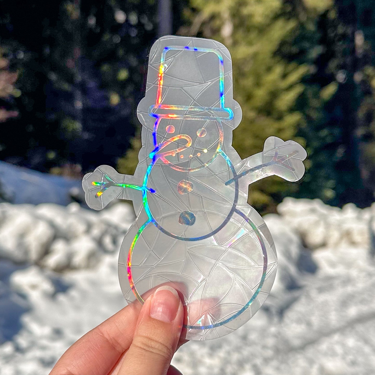 Holographic cozy winter snowman suncatcher cling decal sticker. Snowman has a carrot nose and top hat. Held up in front of snow and trees.