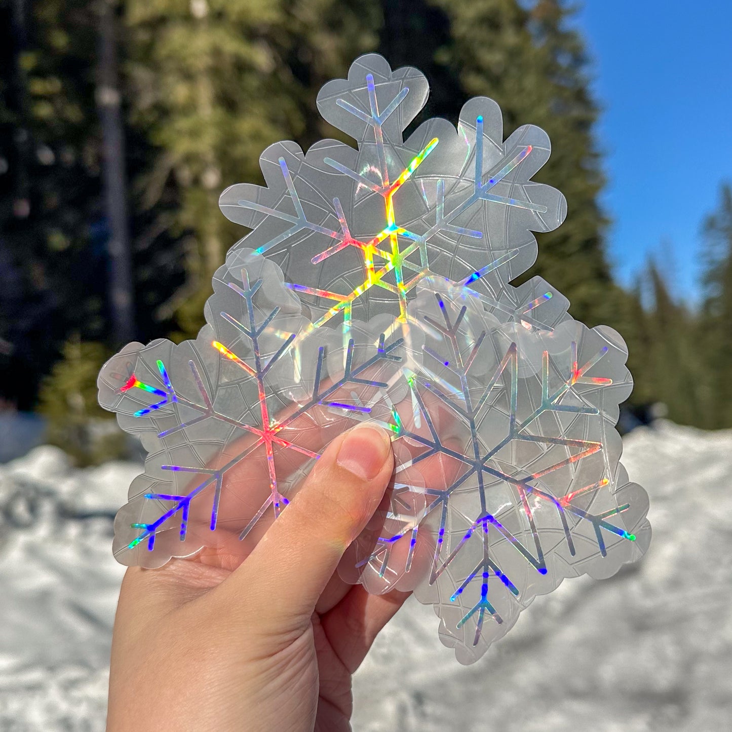 3 Holographic cozy winter snowflake  suncatcher cling decal stickers. 3 Holographic snowflake designs. Held up in front of snow and trees.