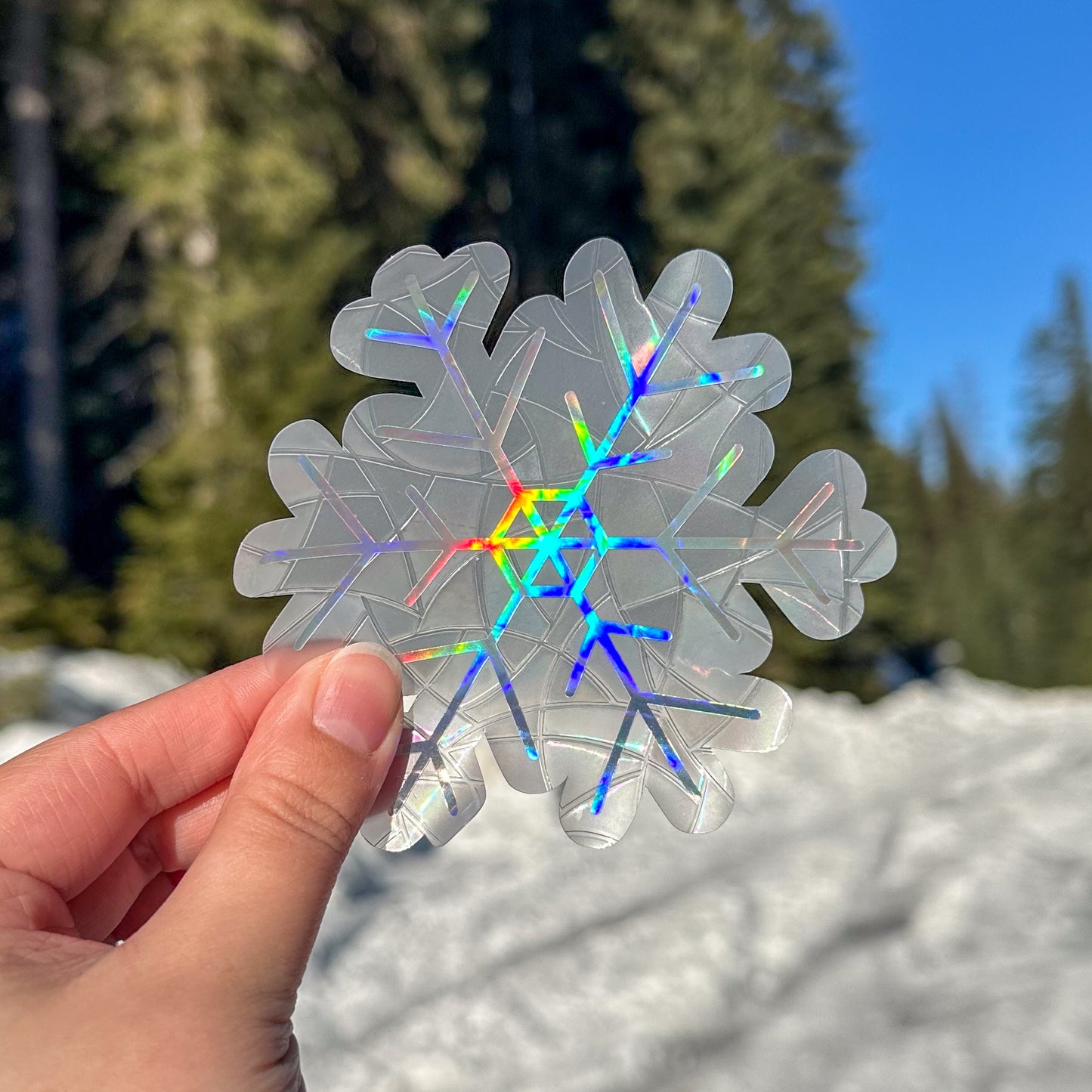 Holographic cozy winter snowflake  suncatcher cling decal sticker. Held up in front of snow and trees.