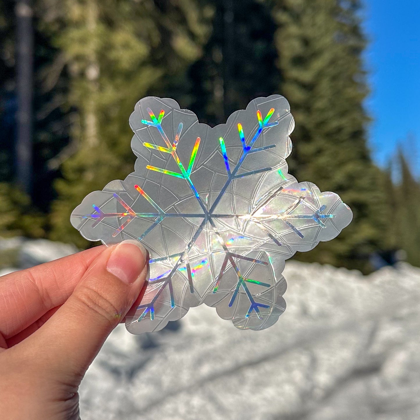 Holographic cozy winter snowflake  suncatcher cling decal sticker. Held up in front of snow and trees.