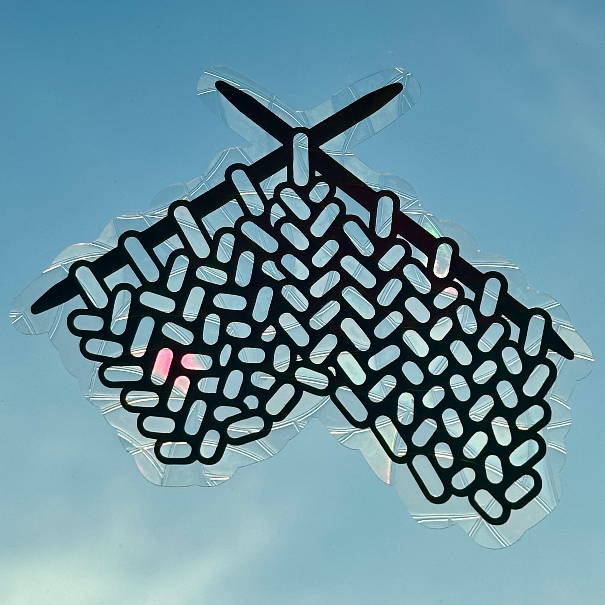 A Knitting Sun Catcher decal on glass in front of a blue sky. 
The design features a knit swatch hanging off of two knitting needles. The design is shiny and holographic, and sits on top of the clear rainbow-making film.