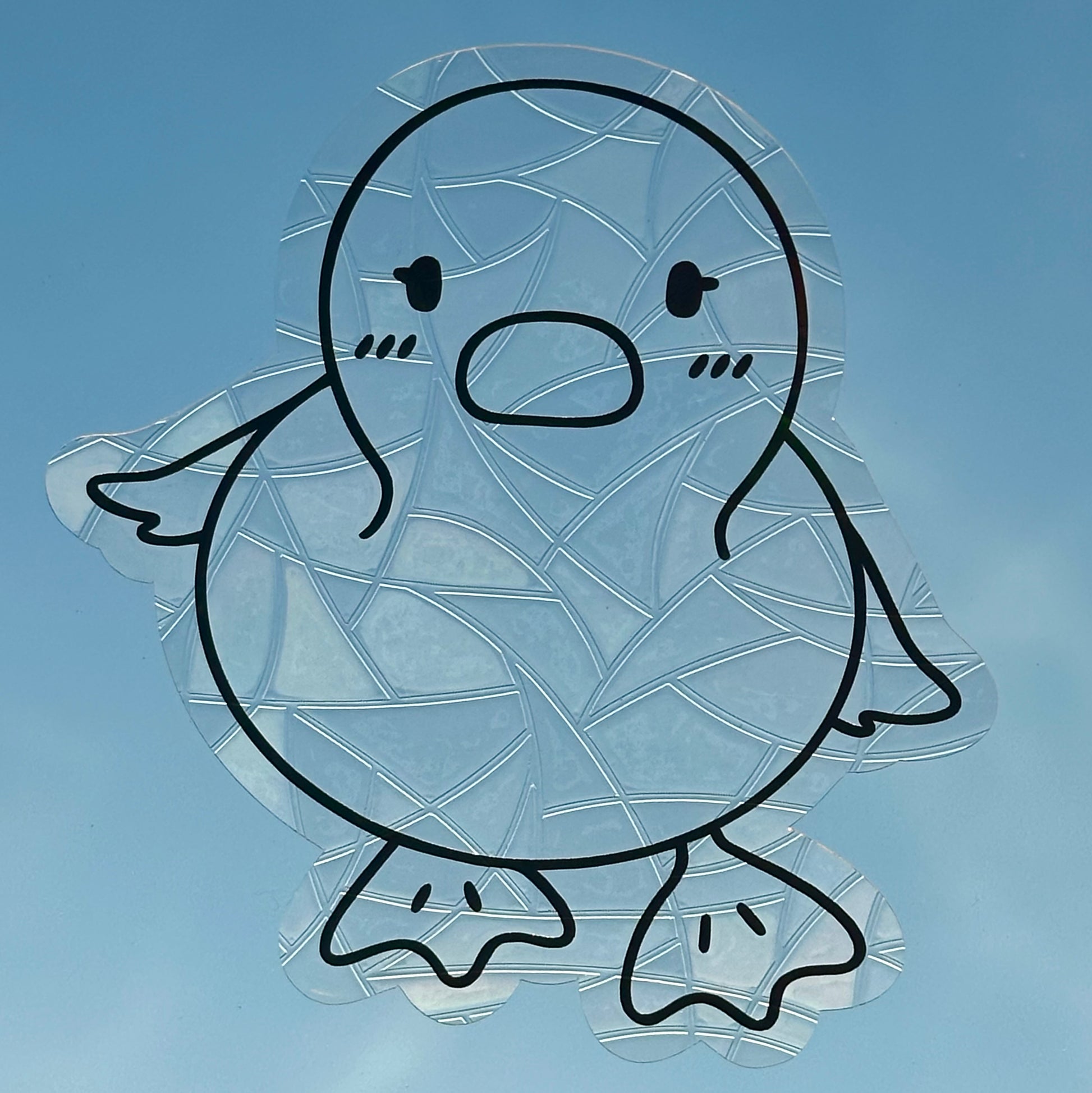 A Sun Catcher decal on glass in front of a blue sky. 
The design features a baby duckling. The design is shiny and holographic, and sits on top of the clear rainbow-making film.