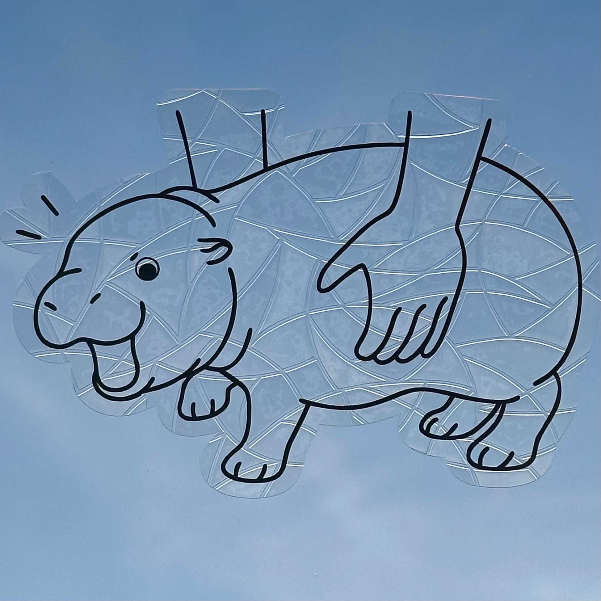 A Sun Catcher decal on glass in front of a blue sky. 
The design features a baby pygmy hippo being picked up. The design is shiny and holographic, and sits on top of the clear rainbow-making film.