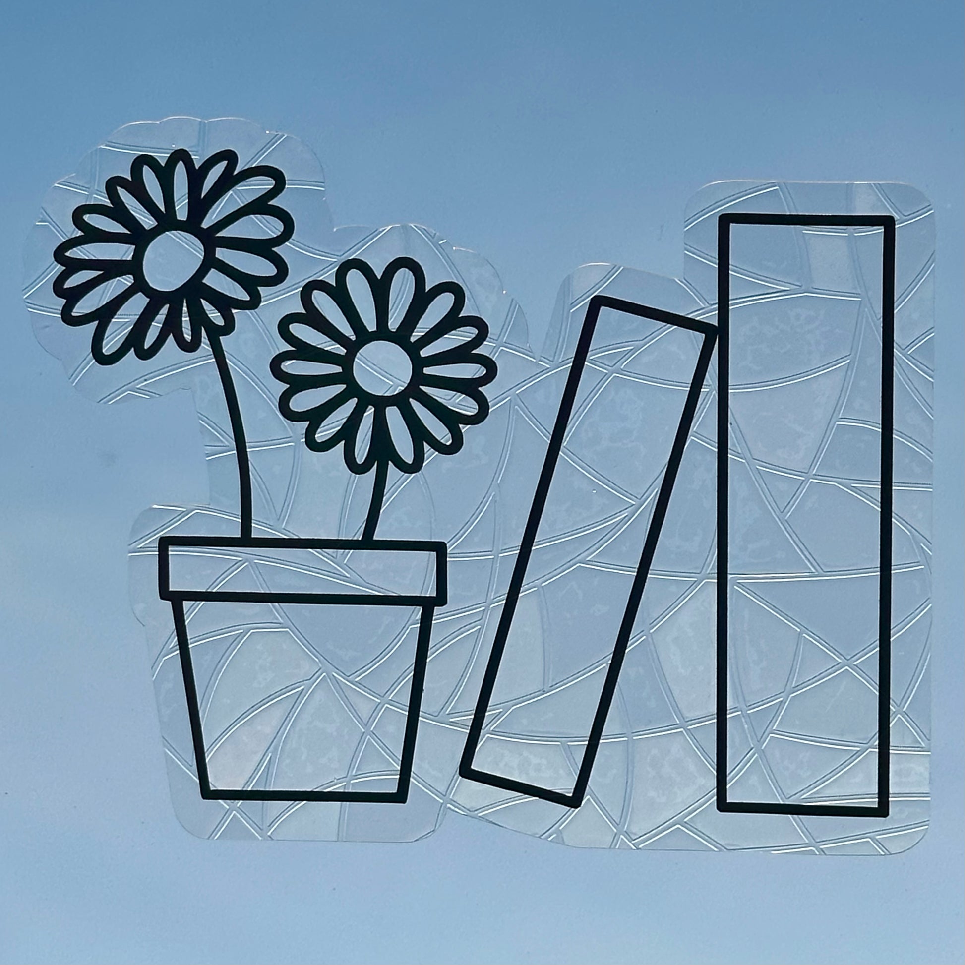 A Sun Catcher decal on glass in front of a blue sky. 
The design features a pot holding two daisies, and next to that, two books. The design is shiny and holographic, and sits on top of the clear rainbow-making film.