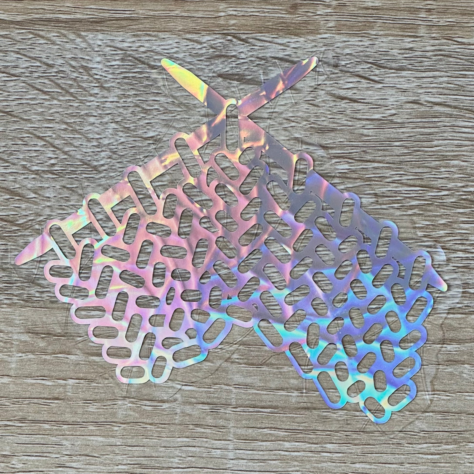 A Knitting Sun Catcher decal on a wooden desk top. 
The design features a knit swatch hanging off of two knitting needles. The design is shiny and holographic, and sits on top of the clear rainbow-making film.