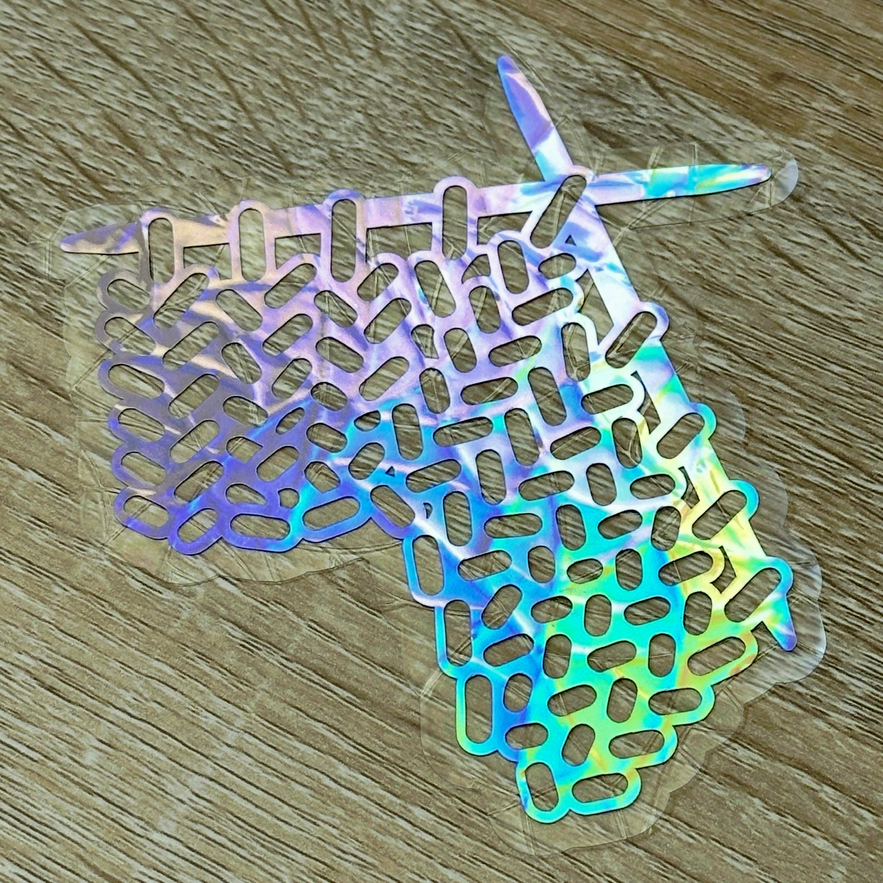 A Knitting Sun Catcher decal on a wooden desk top. 
The design features a knit swatch hanging off of two knitting needles. The design is shiny and holographic, and sits on top of the clear rainbow-making film.