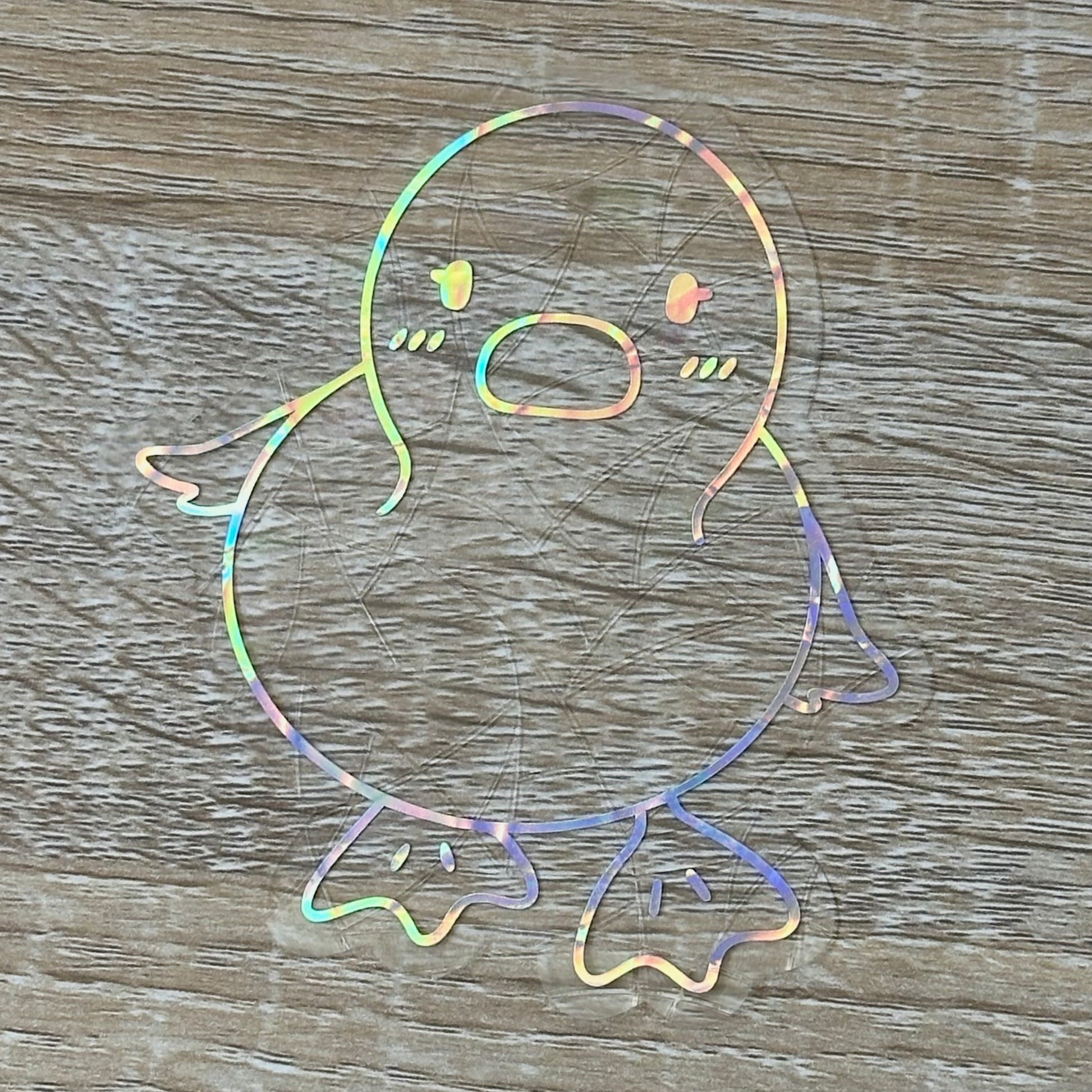 A Sun Catcher decal on a wooden desk top. 
The design features a baby duckling. The design is shiny and holographic, and sits on top of the clear rainbow-making film.