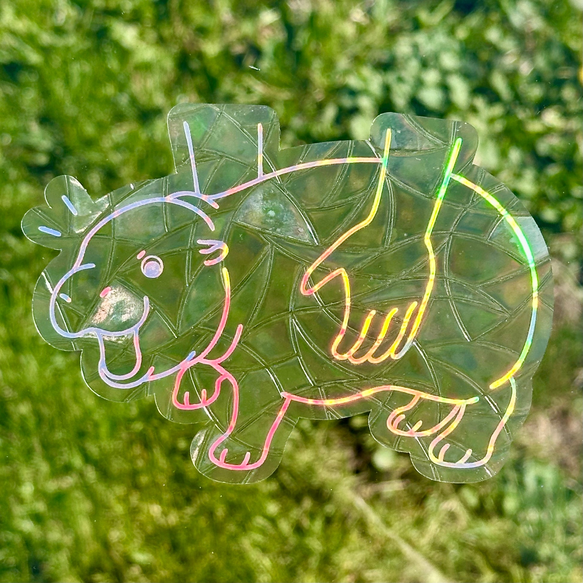 A Sun Catcher decal in front of green grass. 
The design features a baby pygmy hippo being picked up. The design is shiny and holographic, and sits on top of the clear rainbow-making film.