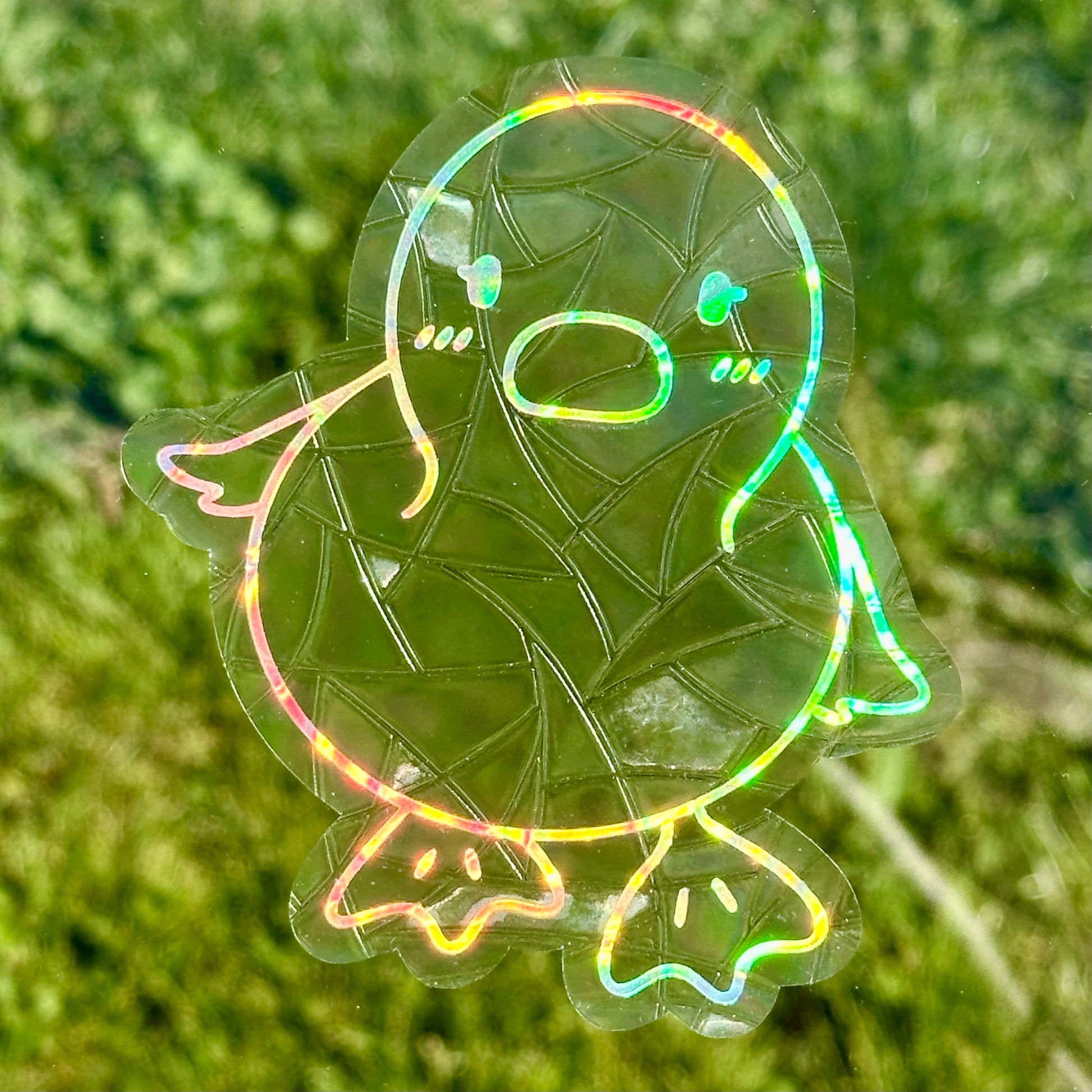 A Sun Catcher decal on glass in front of green grass. 
The design features a baby duckling. The design is shiny and holographic, and sits on top of the clear rainbow-making film.