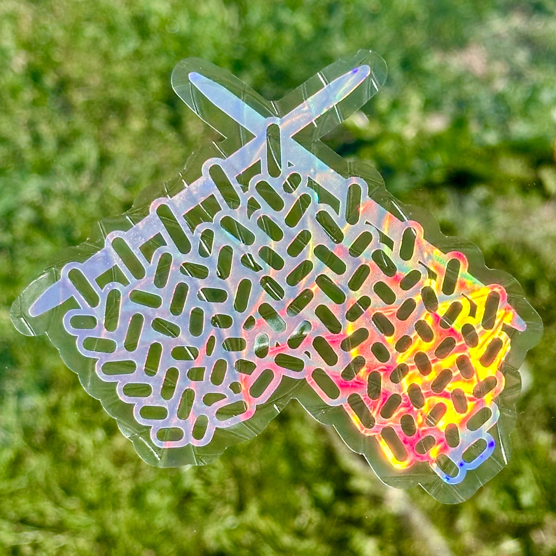 A Knitting Sun Catcher decal in front of green grass. 
The design features a knit swatch hanging off of two knitting needles. The design is shiny and holographic, and sits on top of the clear rainbow-making film.