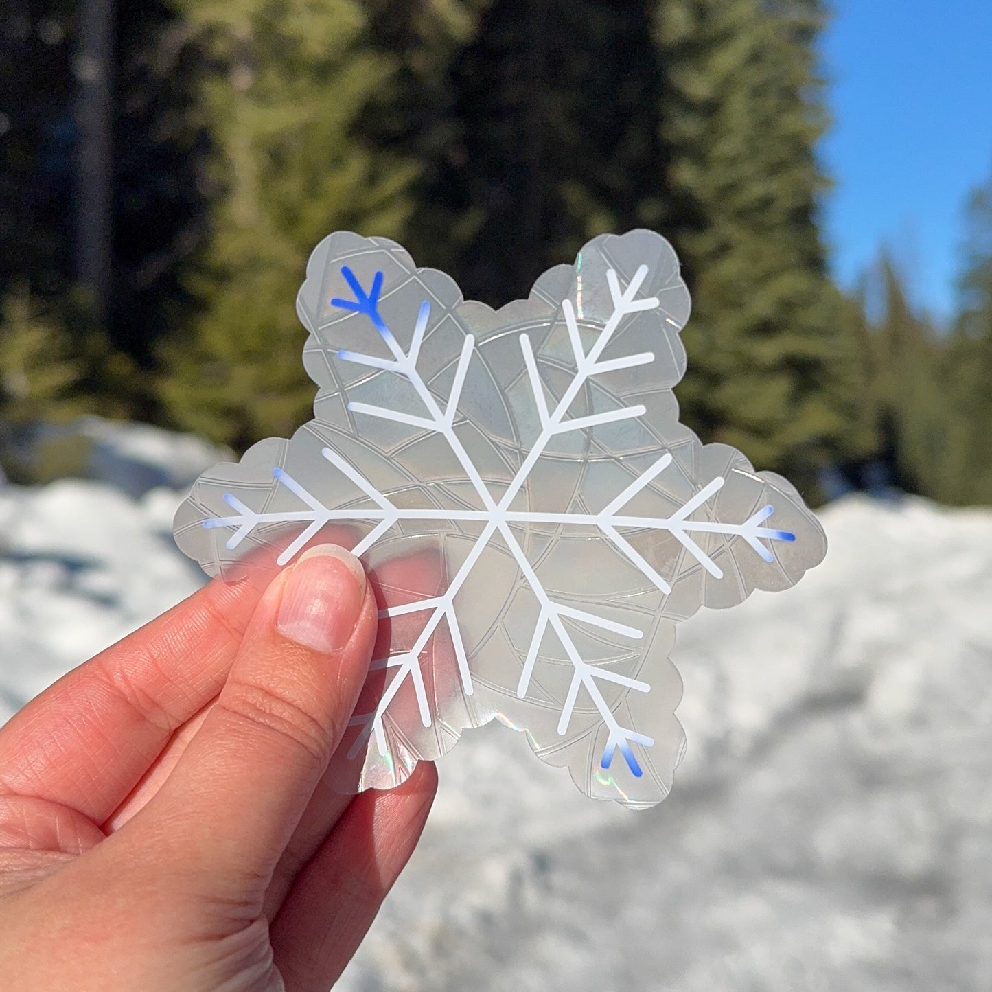 Cozy winter snowflake suncatcher cling decal sticker.
The suncatcher is white, slowly beginning to change color to a deep blue on the edges. Held up in front of snow and trees.