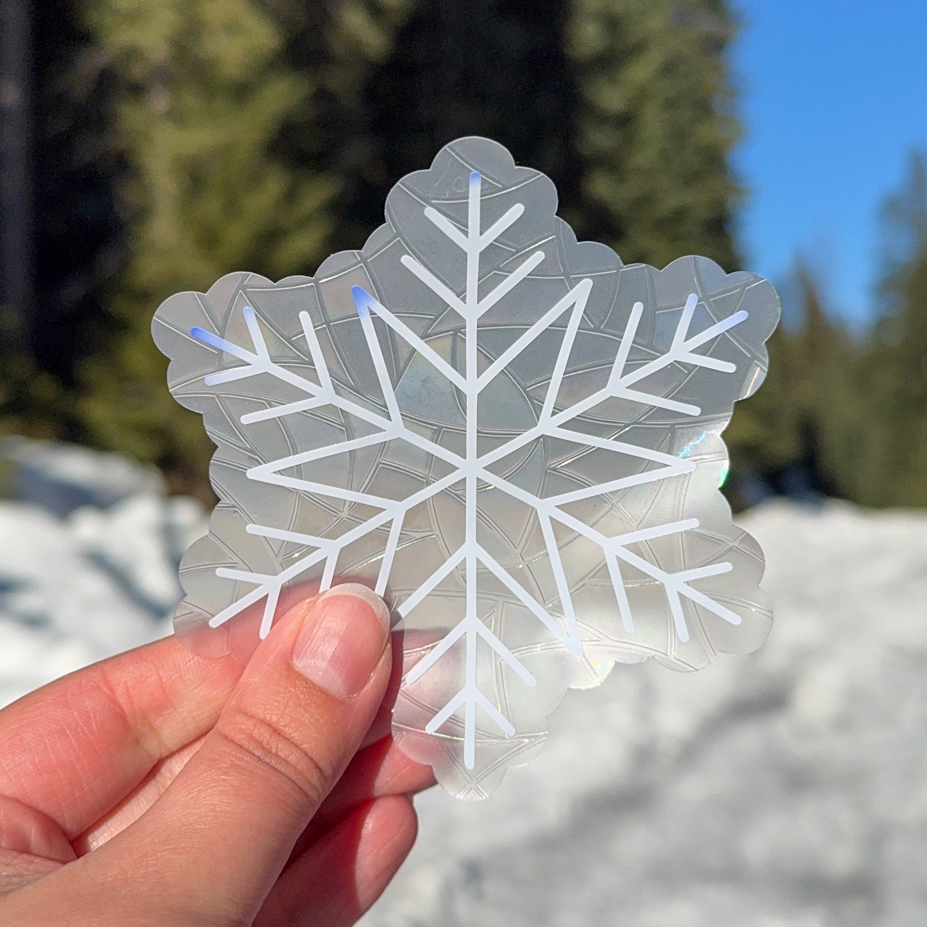 Cozy winter snowflake suncatcher cling decal sticker.
The suncatcher is white, slowly beginning to change color to a deep blue on the edges. Held up in front of snow and trees.