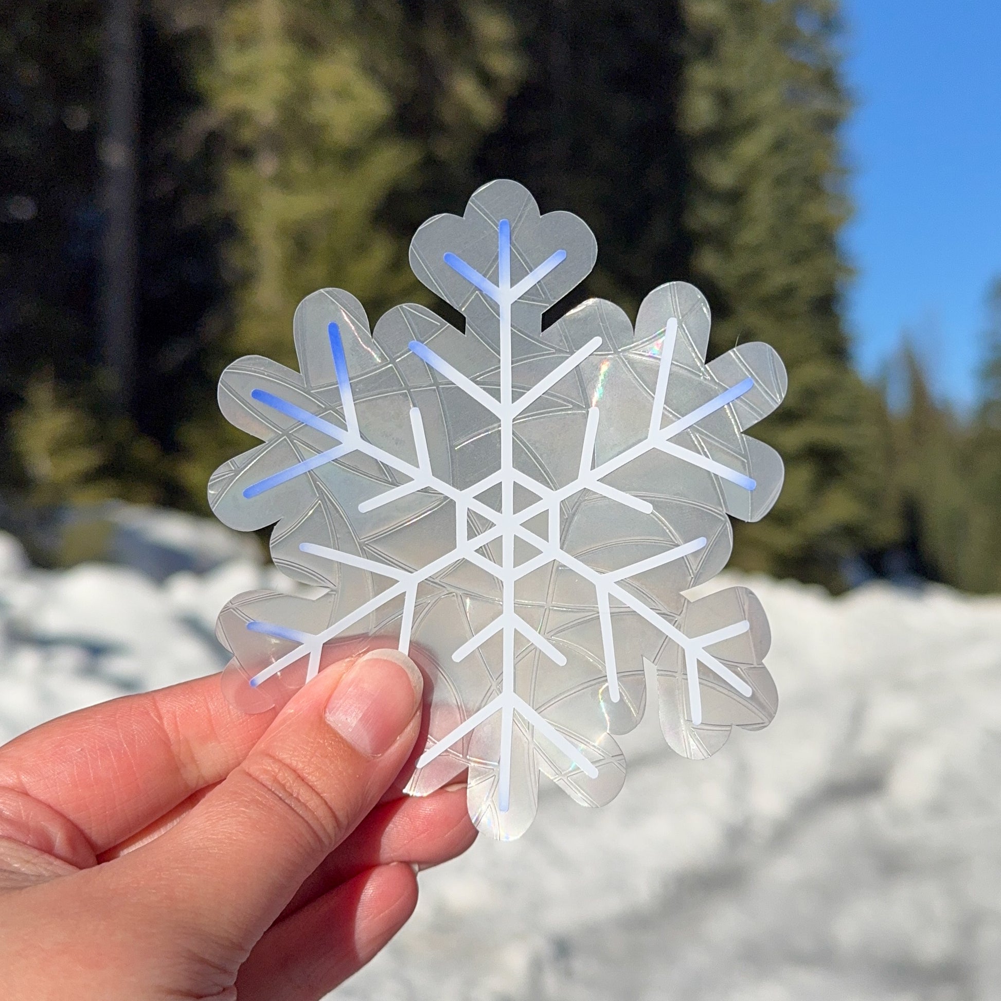 Cozy winter snowflake suncatcher cling decal sticker.
The suncatcher is white, slowly beginning to change color to a deep blue on the edges. Held up in front of snow and trees.