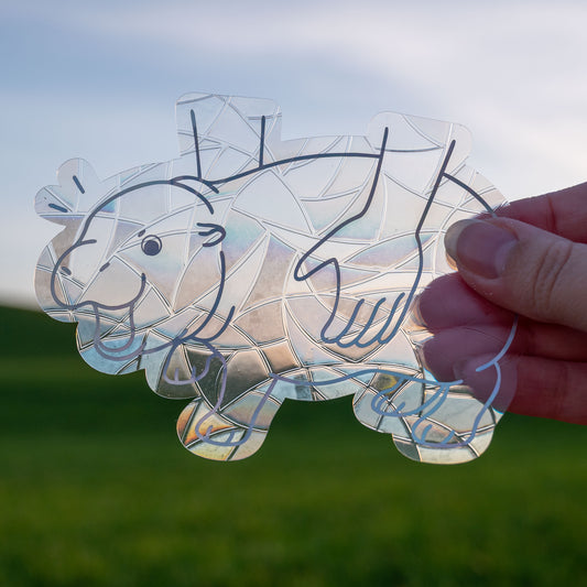 A Sun Catcher decal held up in front of a blue sky and green hills. 
The design features a baby pygmy hippo being picked up. The design is shiny and holographic, and sits on top of the clear rainbow-making film.