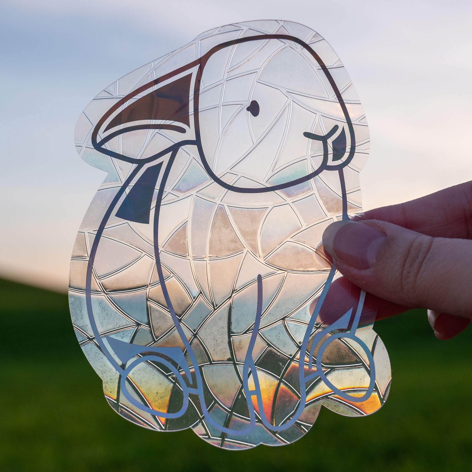 A Sun Catcher decal held up in front of a blue sky and green hills. 
The design features a sitting bunny rabbit. The design is shiny and holographic, and sits on top of the clear rainbow-making film.