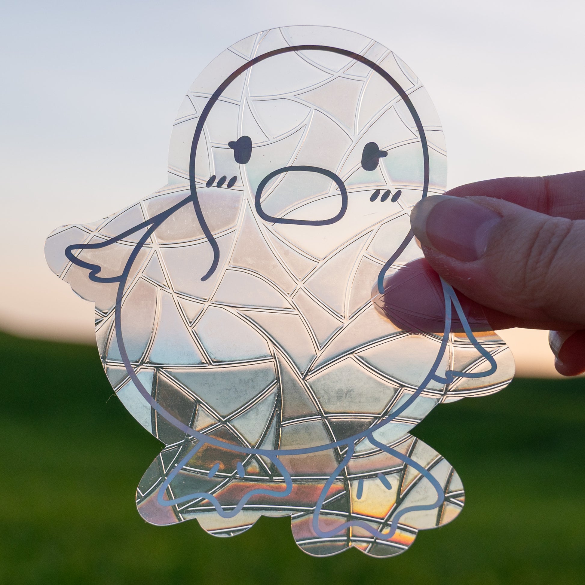 A Sun Catcher decal held up in front of a blue sky and green hills. 
The design features a baby duckling. The design is shiny and holographic, and sits on top of the clear rainbow-making film.