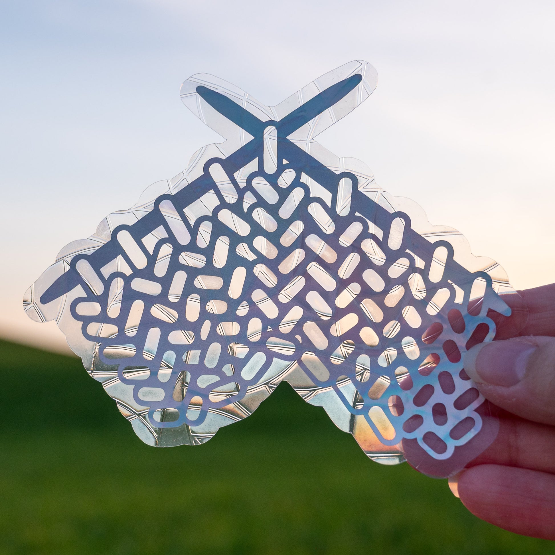 A Knitting Sun Catcher decal held up in front of a blue sky and green hills. 
The design features a knit swatch hanging off of two knitting needles. The design is shiny and holographic, and sits on top of the clear rainbow-making film.