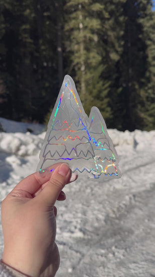 Video of a holographic cozy winter snowy trees suncatcher cling decal sticker. Holographic evergreen trees covered in snow. Held up in front of snow and trees. The protective backing is removed, then it is placed on a window. Then the rainbows that can be made are shown on a wall.