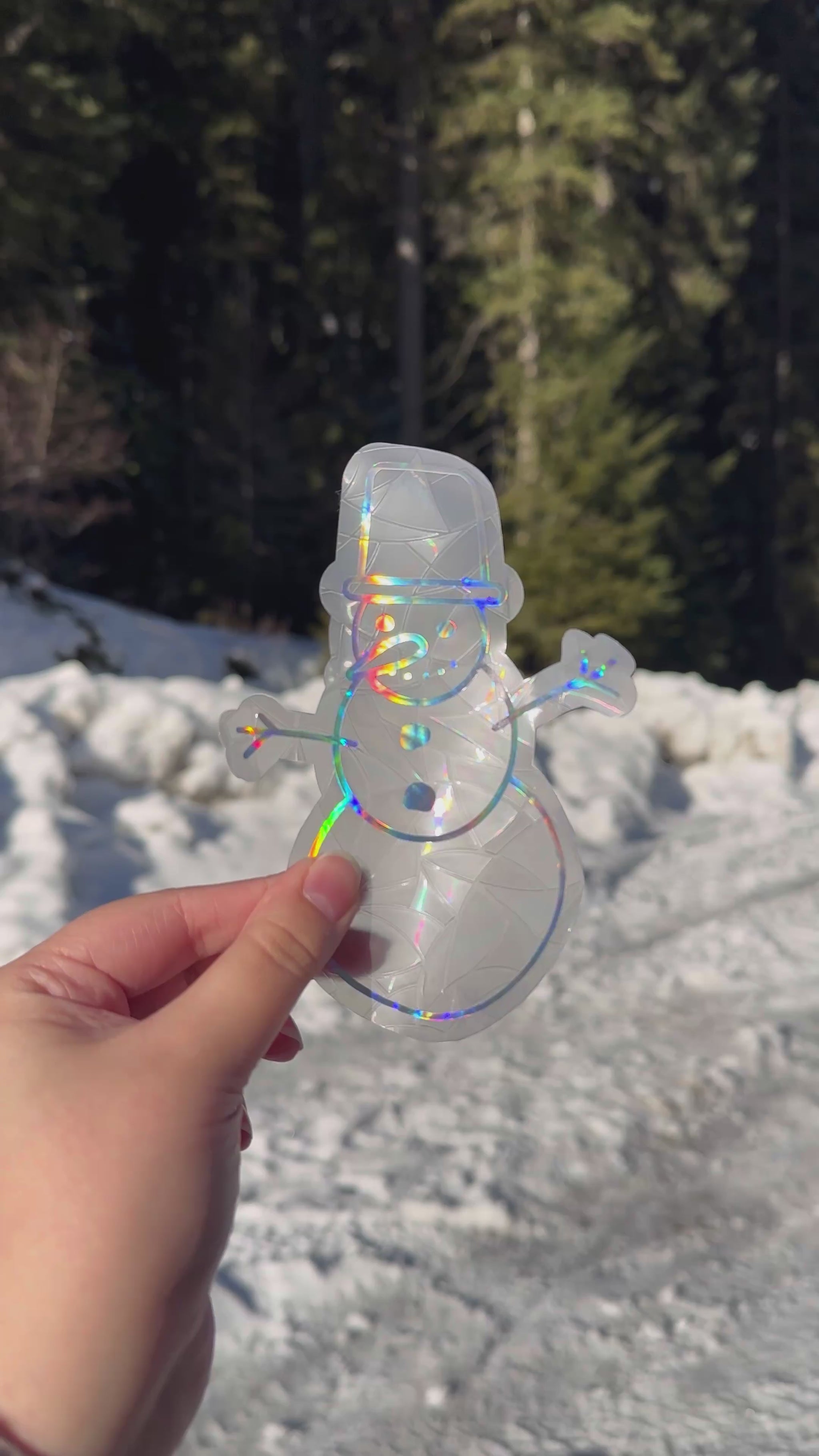 Video of a holographic cozy winter snowman suncatcher cling decal sticker. Holographic snowman with carrot nose and top hat. Held up in front of snow and trees. 
The protective backing is removed, then it is placed on a window. Then the rainbows that can be made are shown on a wall.