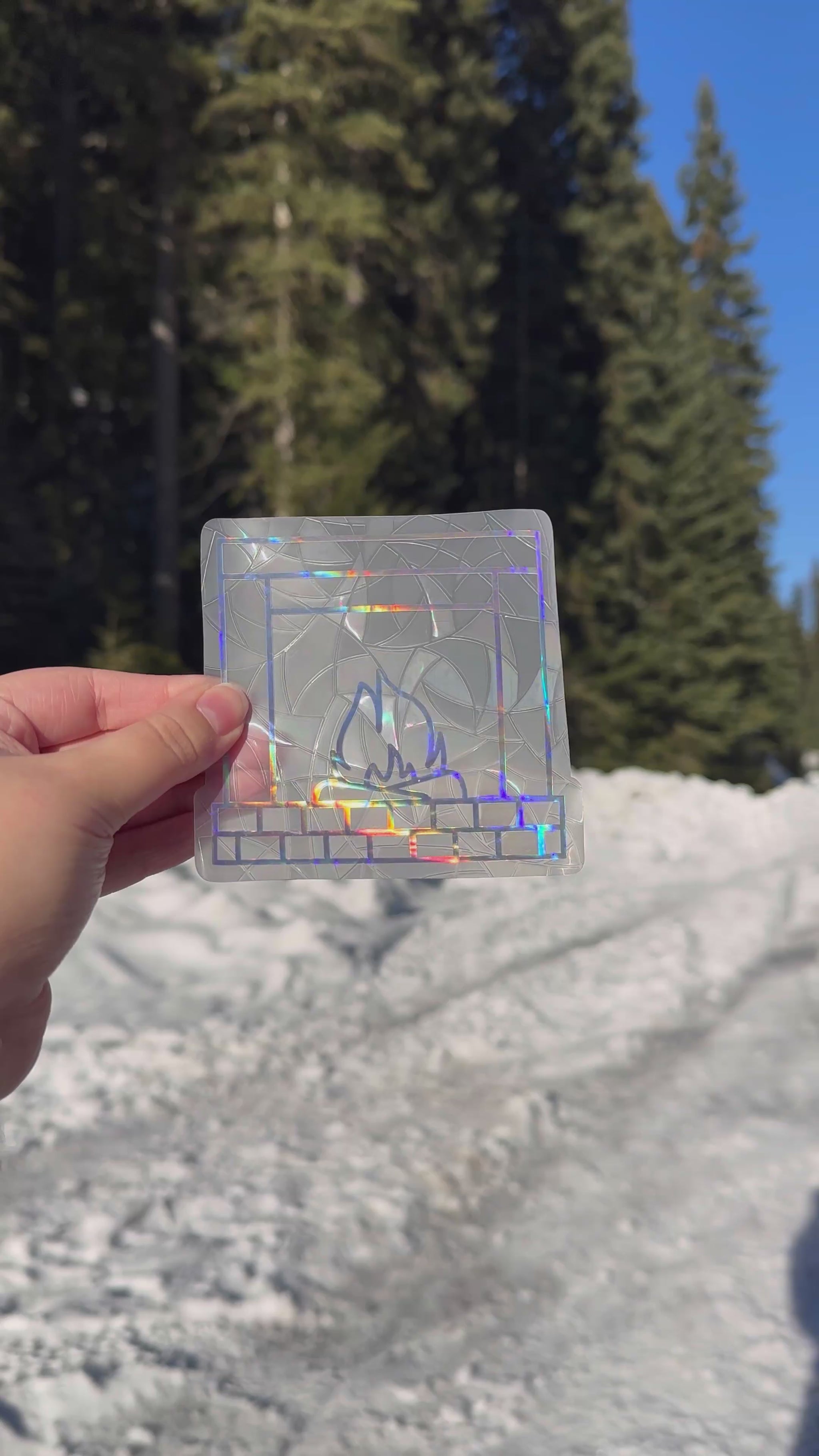 Video of a holographic cozy winter fireplace suncatcher cling decal sticker. Holographic brick fireplace with two logs and a fire in the center. Held up in front of snow and trees. 
The protective backing is removed, then it is placed on a window. Then the rainbows that can be made are shown on a wall.