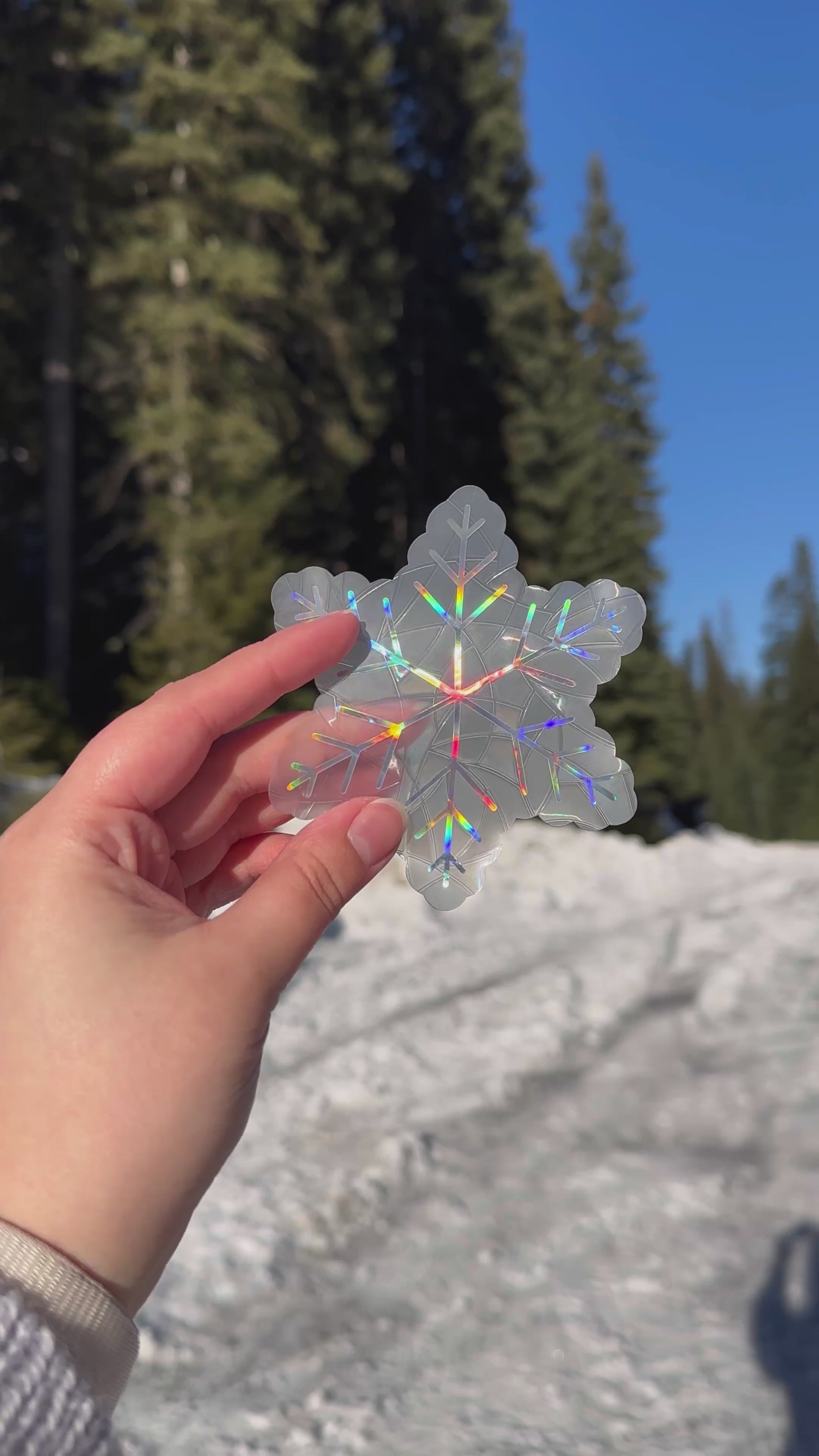 Video of 3 Holographic cozy winter snowflake suncatcher cling decal stickers. 3 Holographic snowflake designs. Held up in front of snow and trees.
The protective backing is removed, then they are placed on a window. Then the rainbows that can be made are shown on a wall.